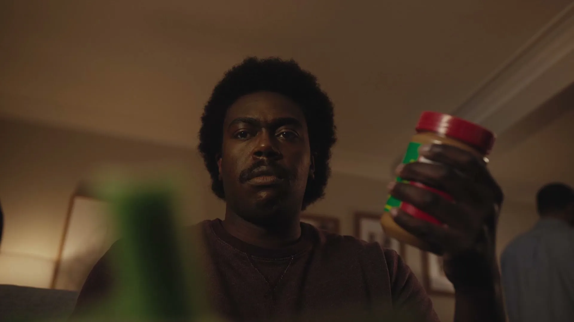 A man with an afro and mustache looks at a jar of peanut butter in his hand, with a blurry green object in the foreground.