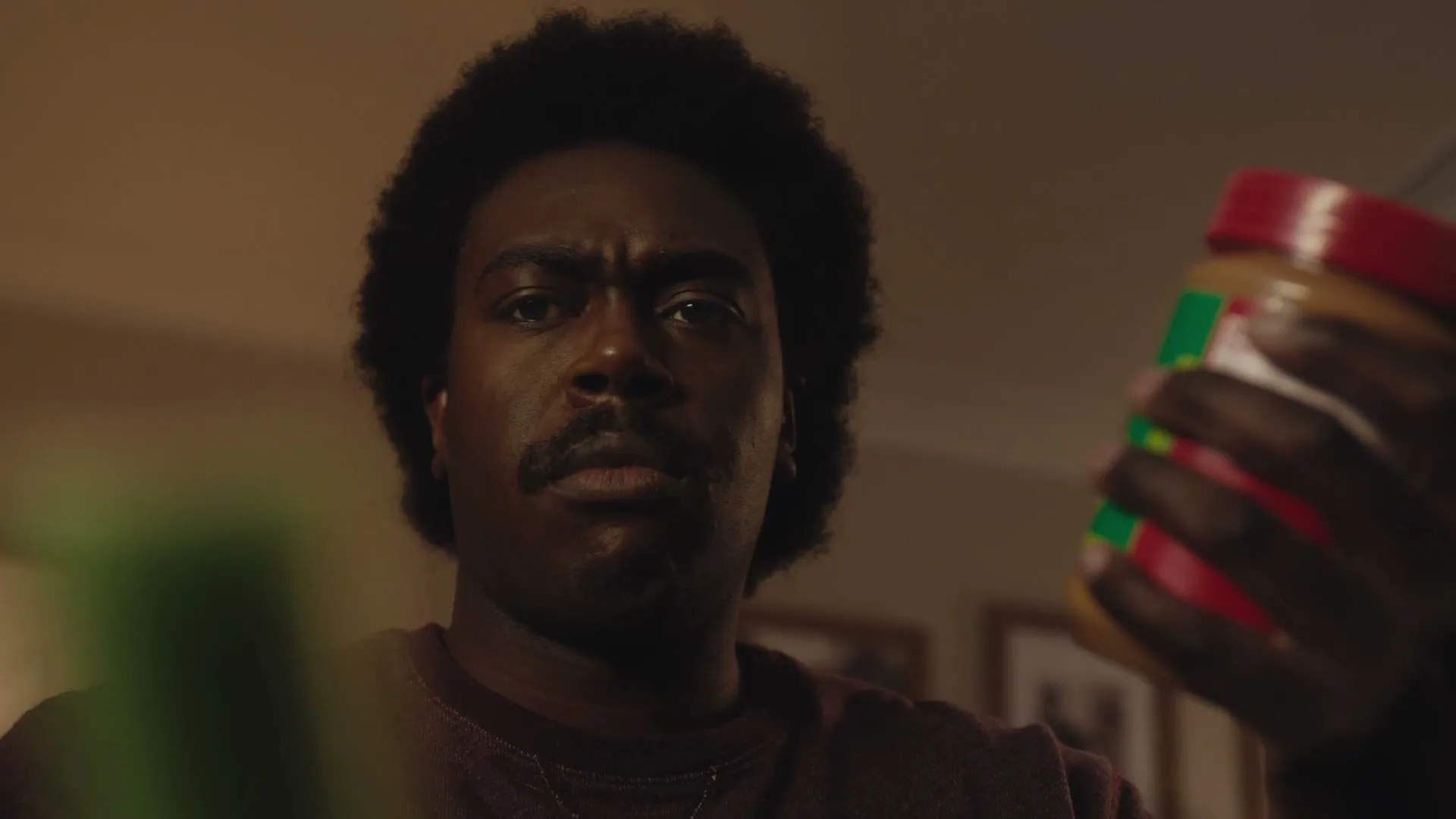 A Black man with an afro and mustache looks intently at a jar of Jif peanut butter in his hand, in a dimly lit interior.