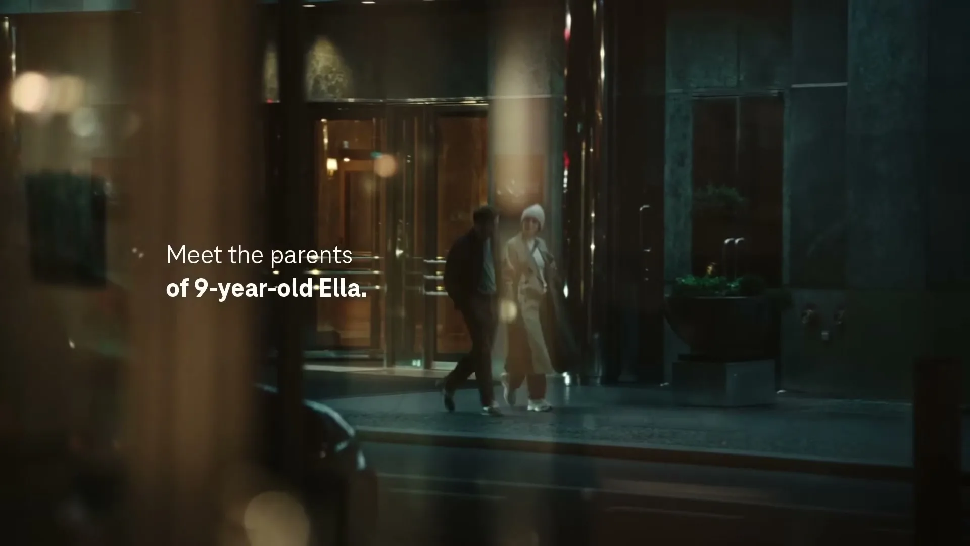 A man and a woman, dressed in casual attire, walk out of a building and onto a city sidewalk at night.