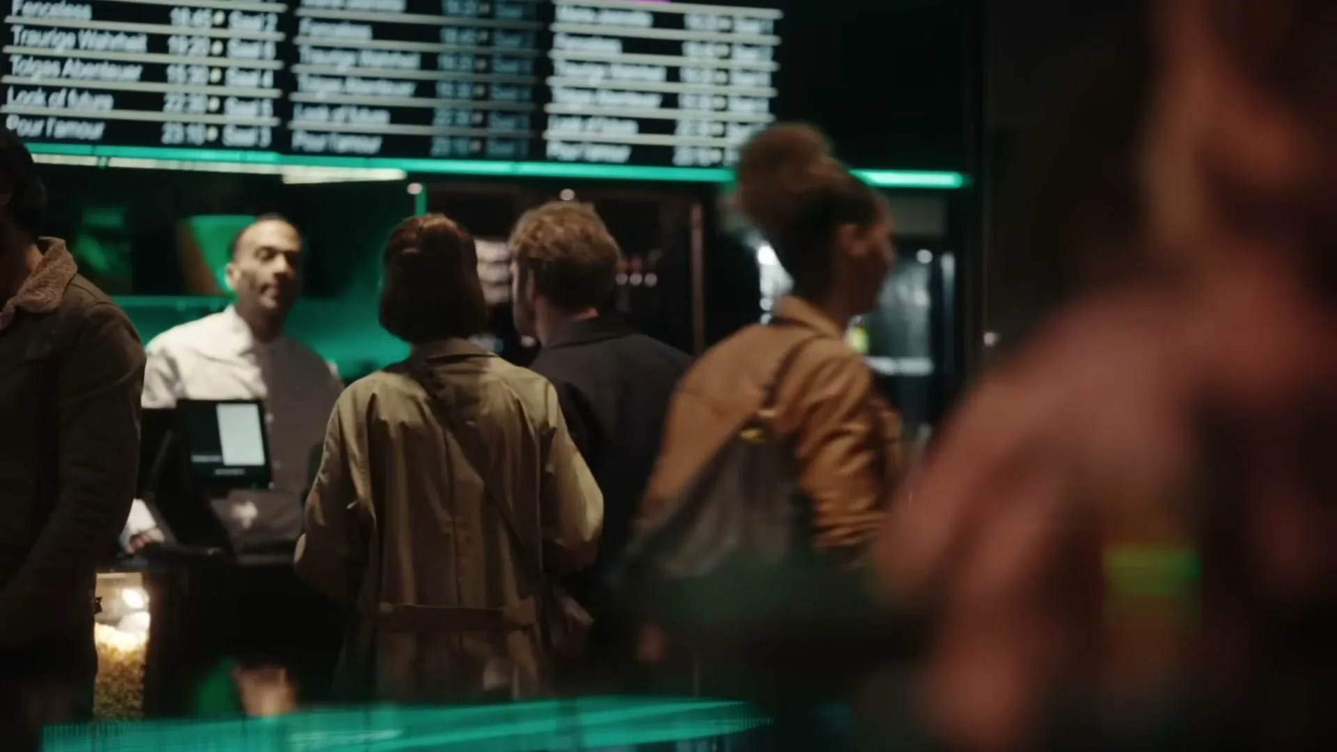 A diverse group of people stand in line at a concession stand, with a man in a white shirt serving customers in a dimly lit cinema lobby.