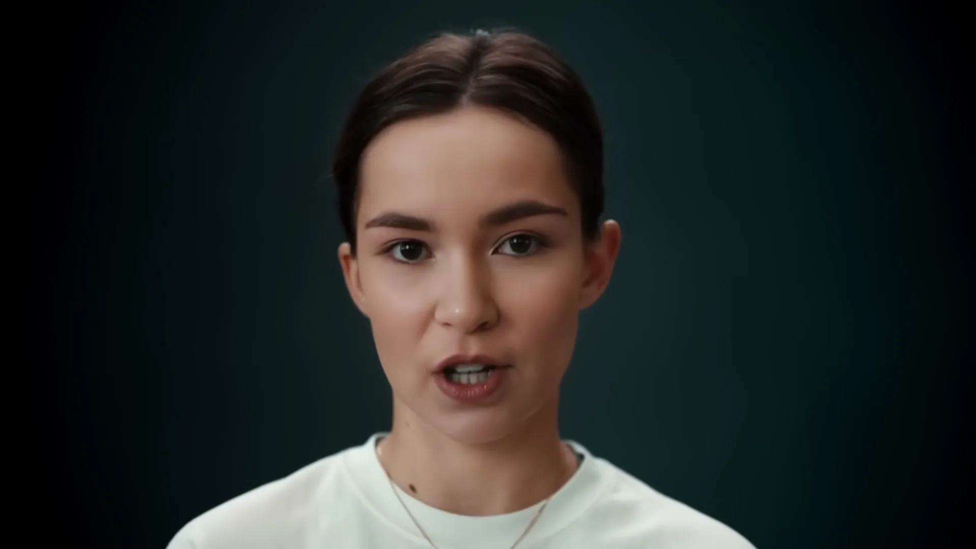 A young woman with dark hair and a white t-shirt speaks directly to the camera against a dark blue background.