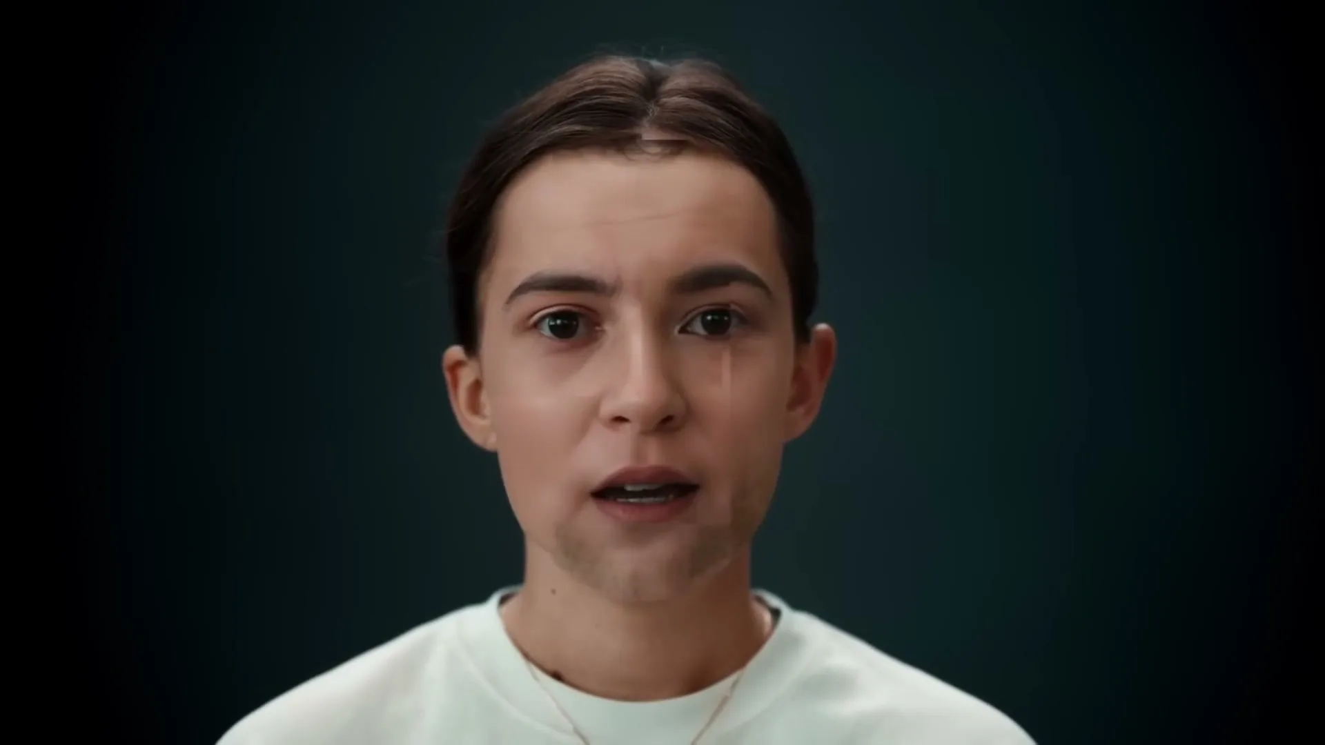 A young woman with dark hair and a white t-shirt speaks directly to the camera, her face digitally altered to appear as a young man, against a dark background.