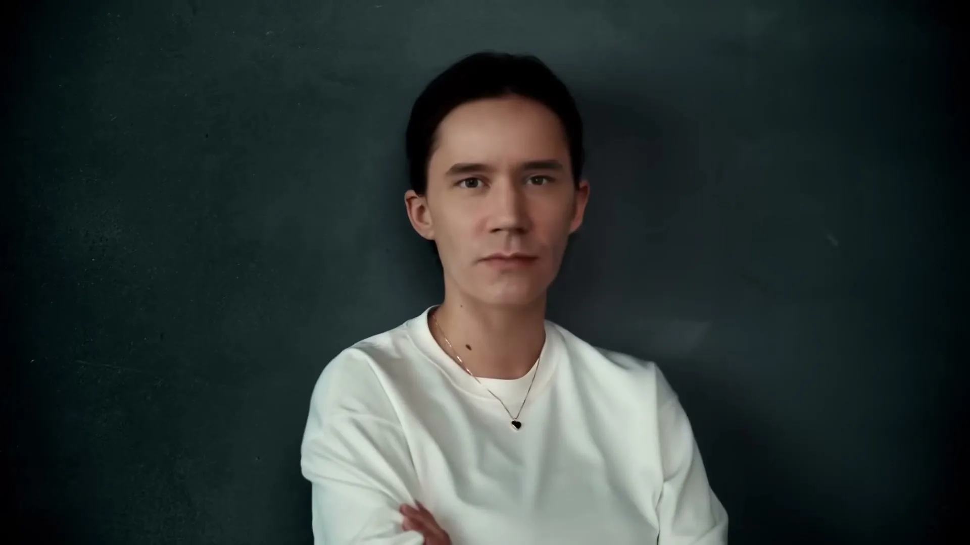 A young person with dark hair and a white shirt stands against a dark gray wall, looking directly at the camera with a serious expression.