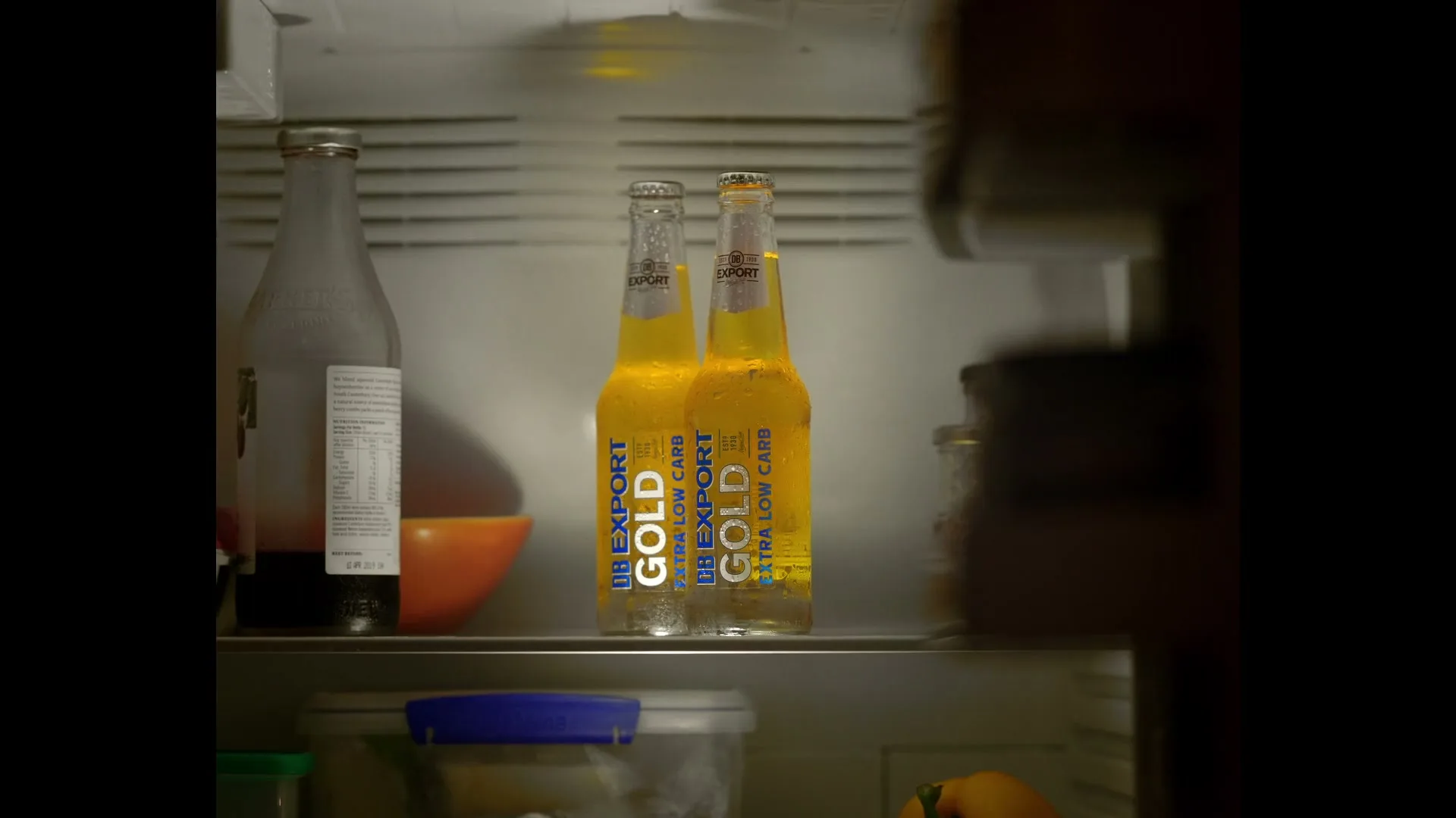 A hand reaches into a refrigerator to grab a bottle of DB Export Gold beer, with another bottle remaining inside.