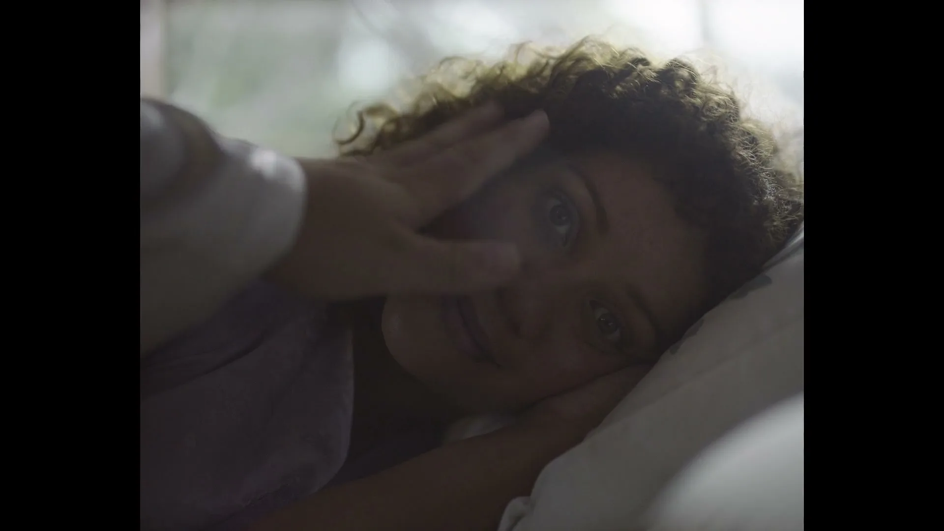 A woman with curly hair lies in bed, smiling softly as a hand gently caresses her cheek, in a dimly lit bedroom.