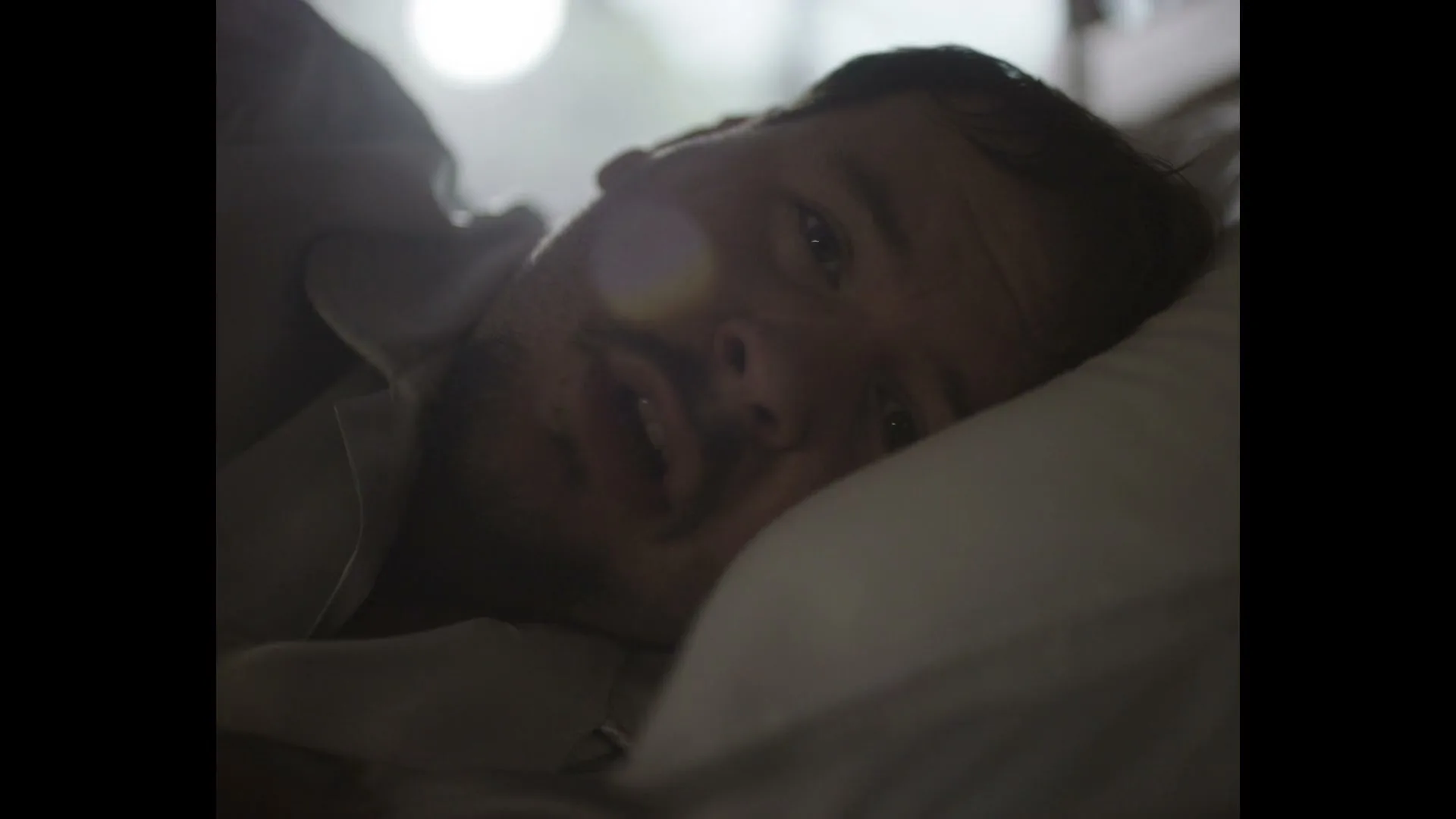 A man in his 30s lies in bed, looking tired and slightly confused, as a faint, ghostly overlay of a kitchen scene appears over him.