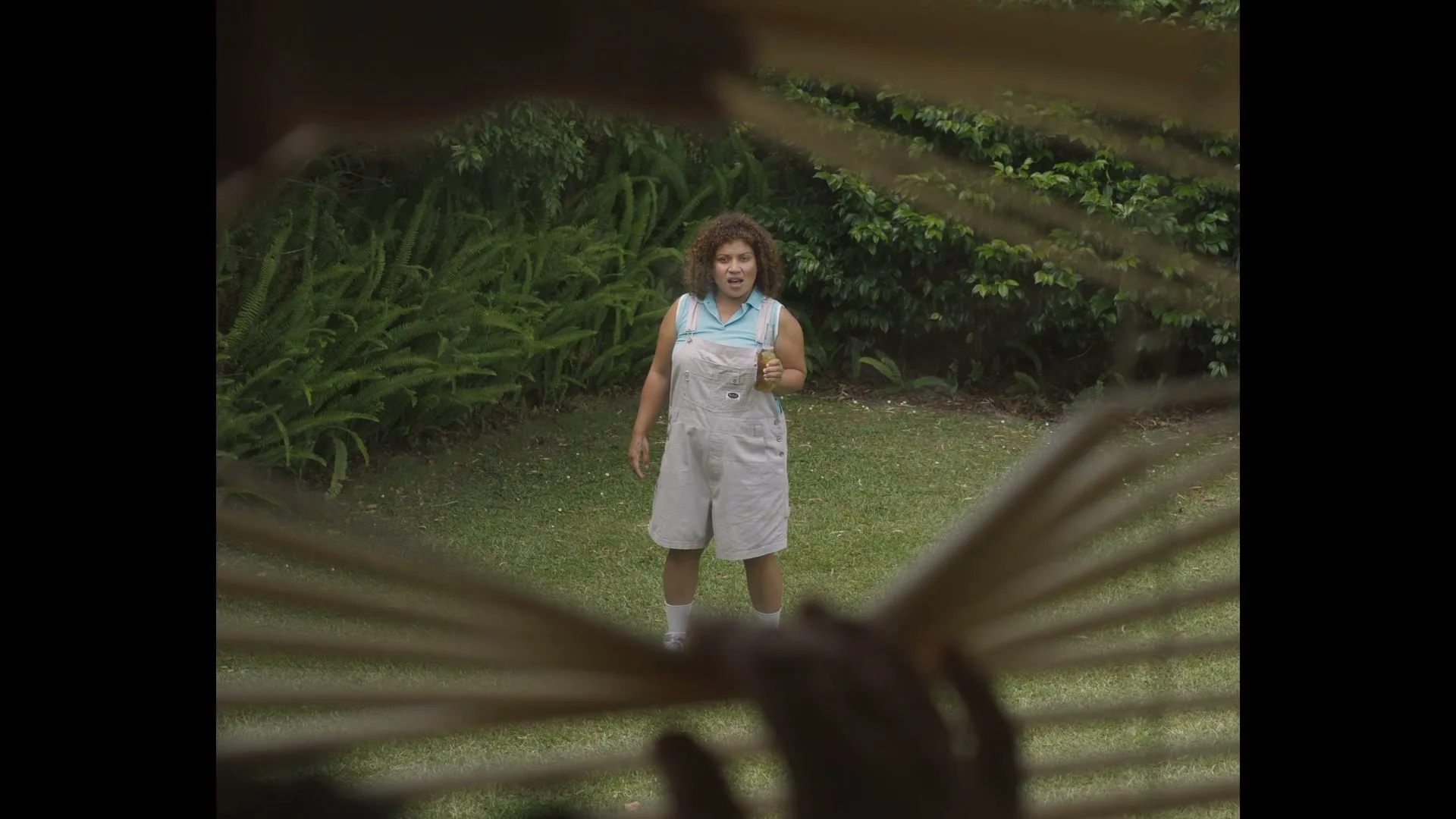 A woman with curly hair in overalls stands in a grassy backyard, holding a beer bottle, seen through parted window blinds.