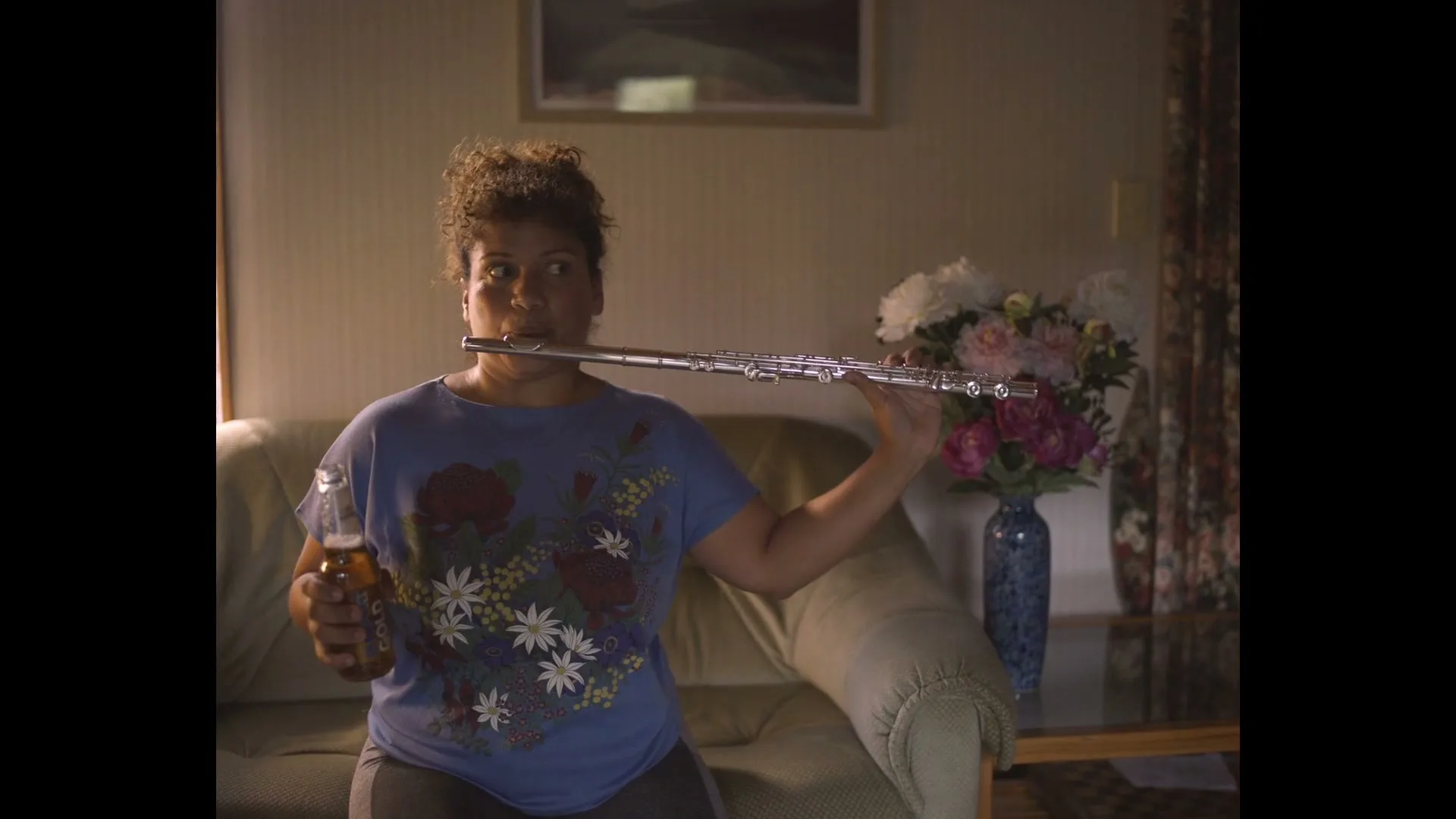 A man sits on a couch holding a beer, then a woman with curly hair plays the flute while holding a beer in a dimly lit living room.