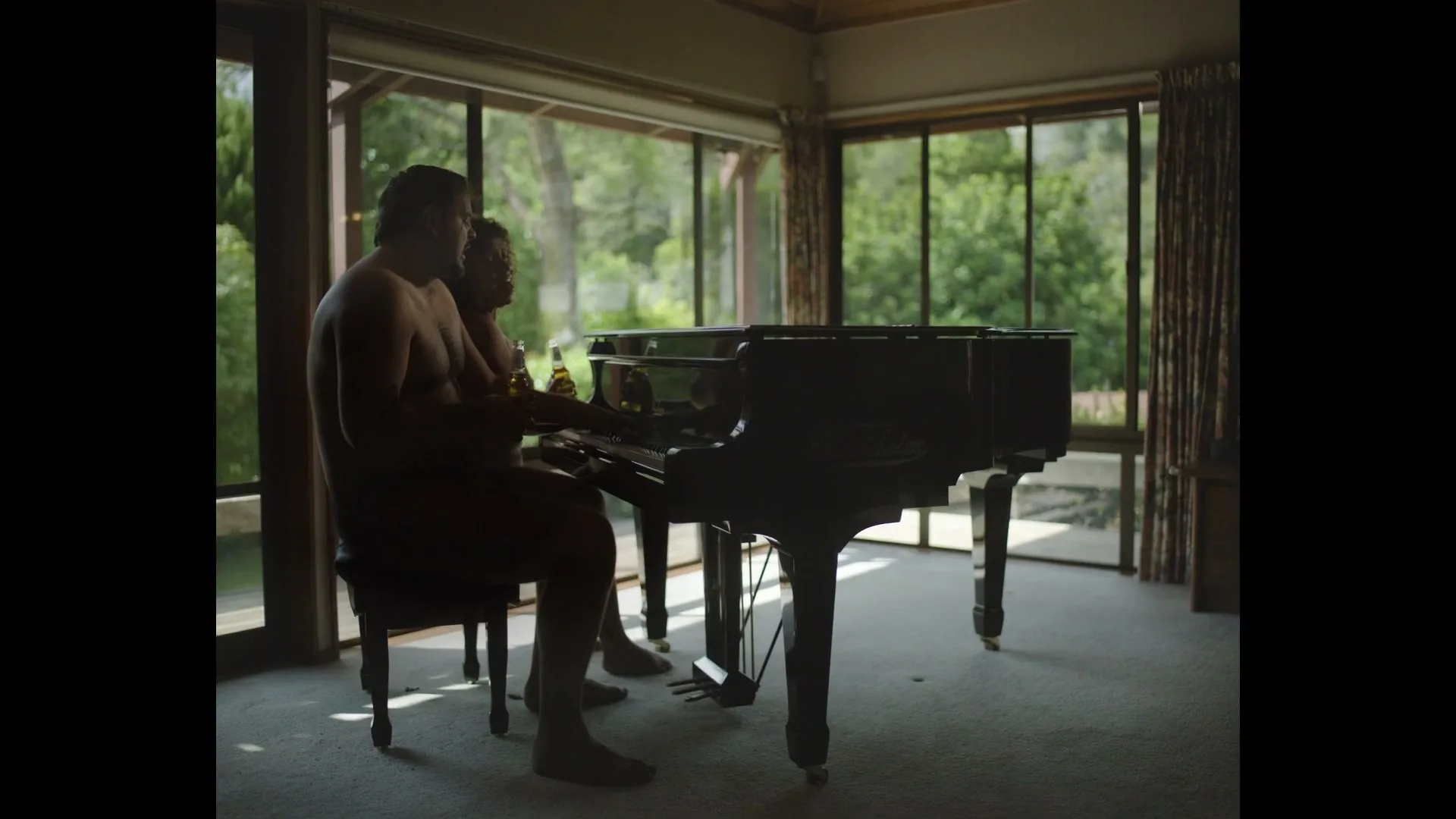Two naked men, one with a beer, play piano together in a sunlit room with large windows overlooking a lush green exterior.
