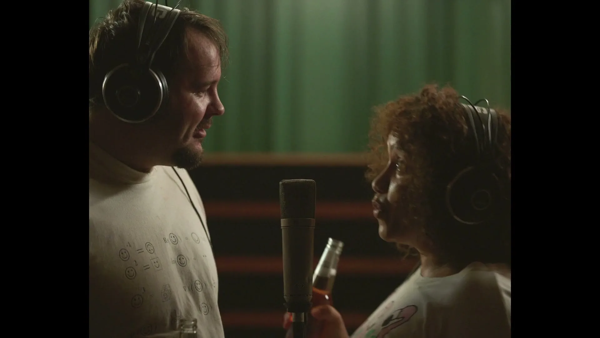 A man and a woman, both wearing headphones, stand close to a microphone in a recording studio, smiling at each other while holding bottles.