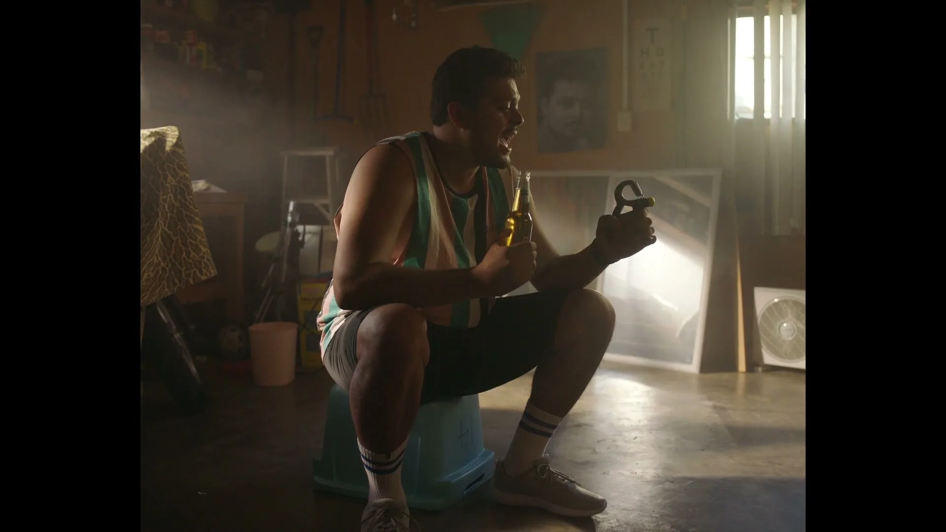 A man in his 30s, wearing a tank top and shorts, sits on a blue stool in a garage, holding a beer bottle and a hand gripper, looking thoughtful.