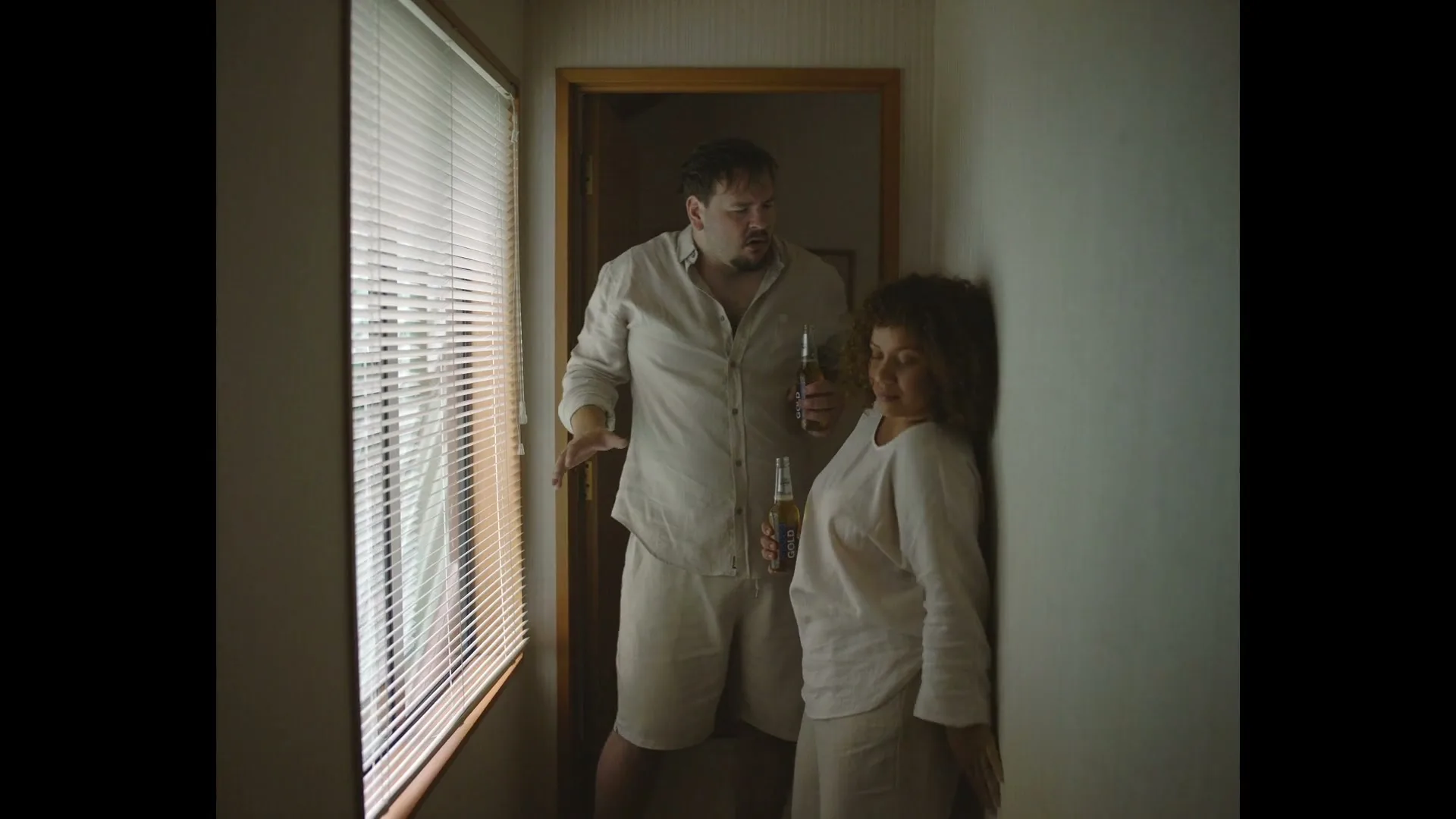 A man and a woman, both wearing light-colored loungewear, stand in a hallway holding beer bottles, looking out a window with blinds.