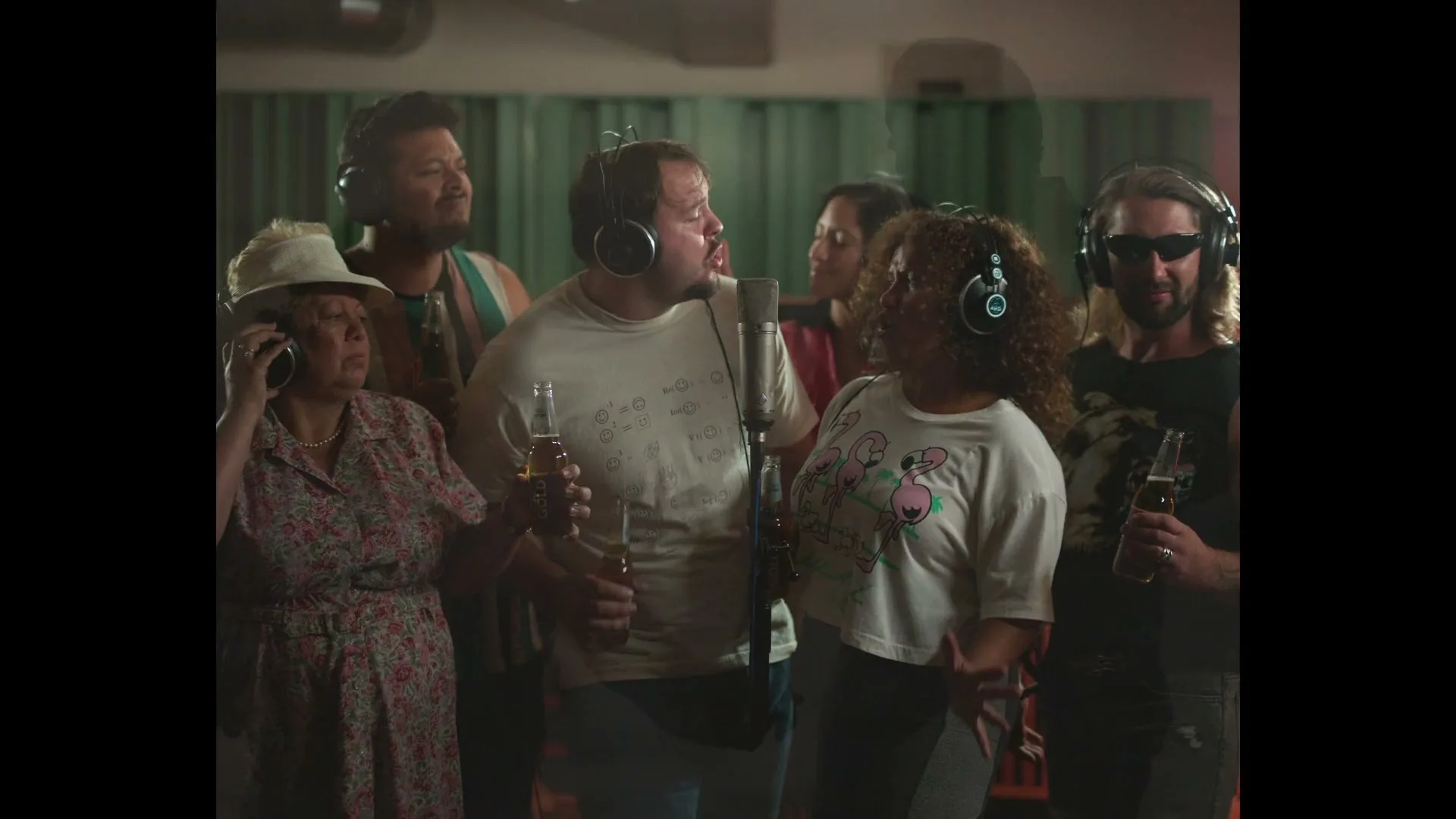A diverse group of six people, wearing headphones and holding beer bottles, sing passionately into a microphone in a recording studio.
