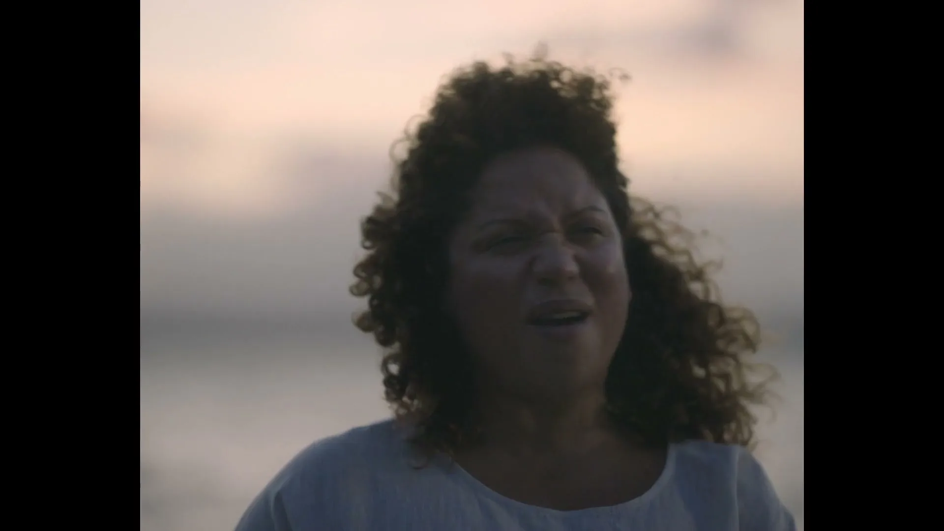 A woman with curly hair, wearing a light-colored top, stands outdoors at dusk, looking around with a slightly confused expression.