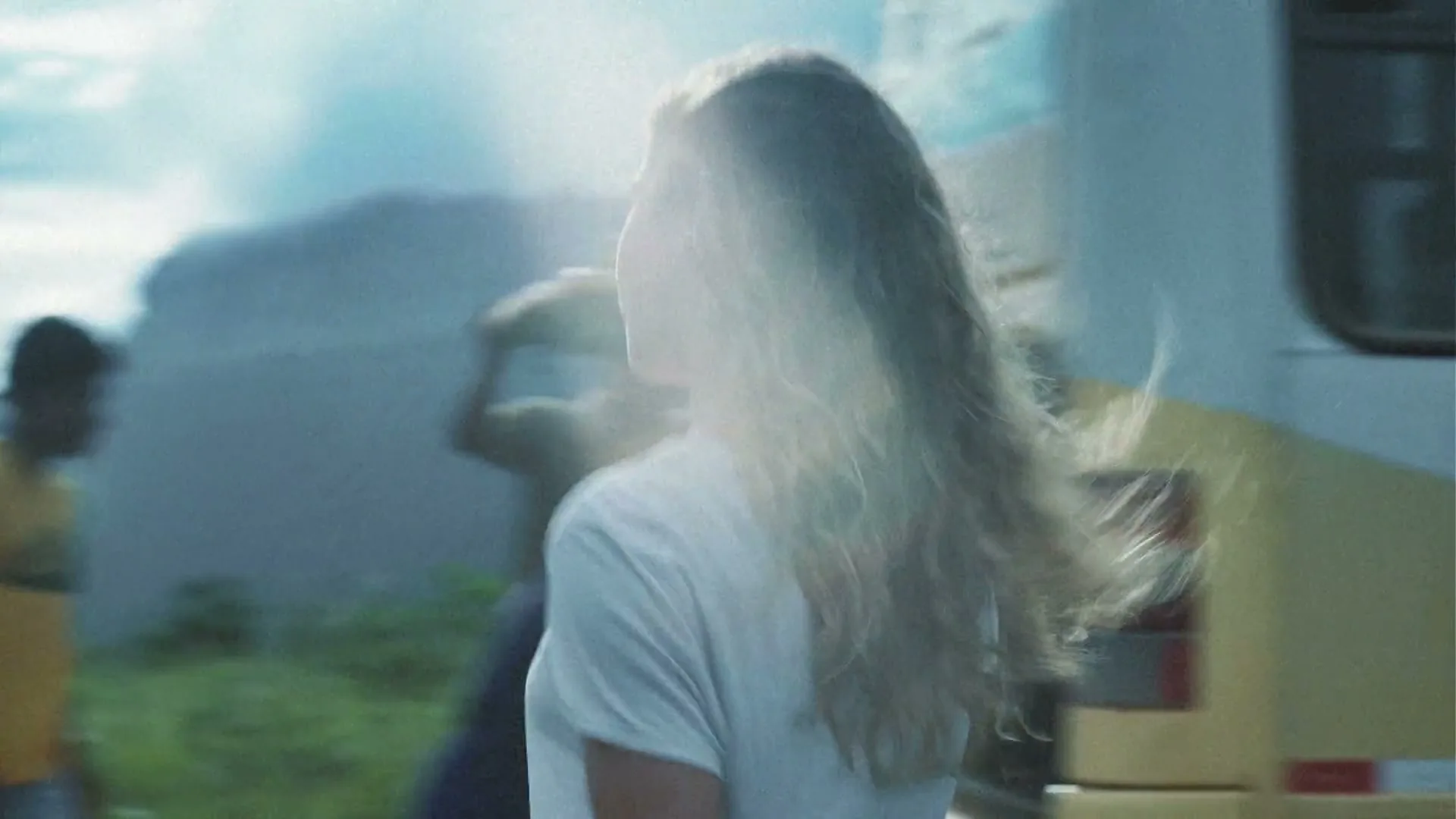 A young woman with long, wavy hair smiles and looks around, then turns to walk with friends towards a large, flat-topped mountain under a cloudy sky.