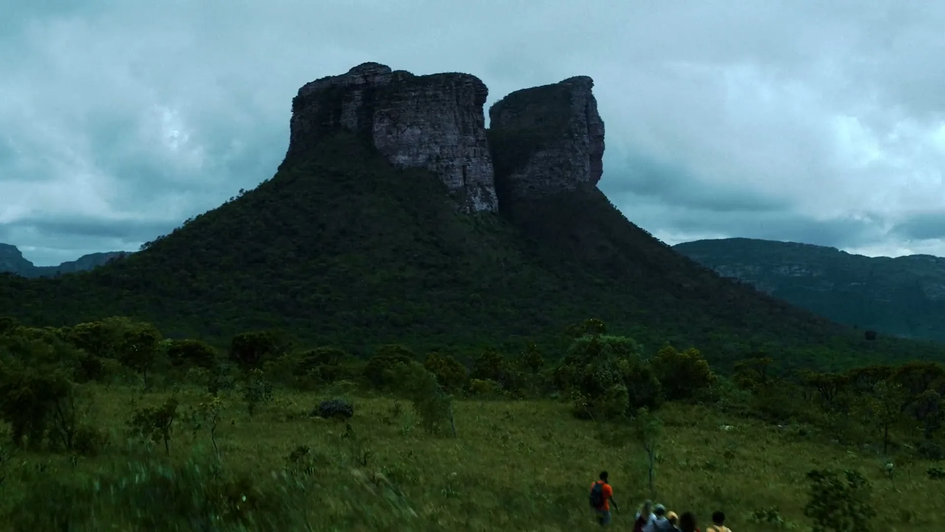 A group of four hikers walk through a grassy field towards two large, flat-topped mountains under a cloudy sky.