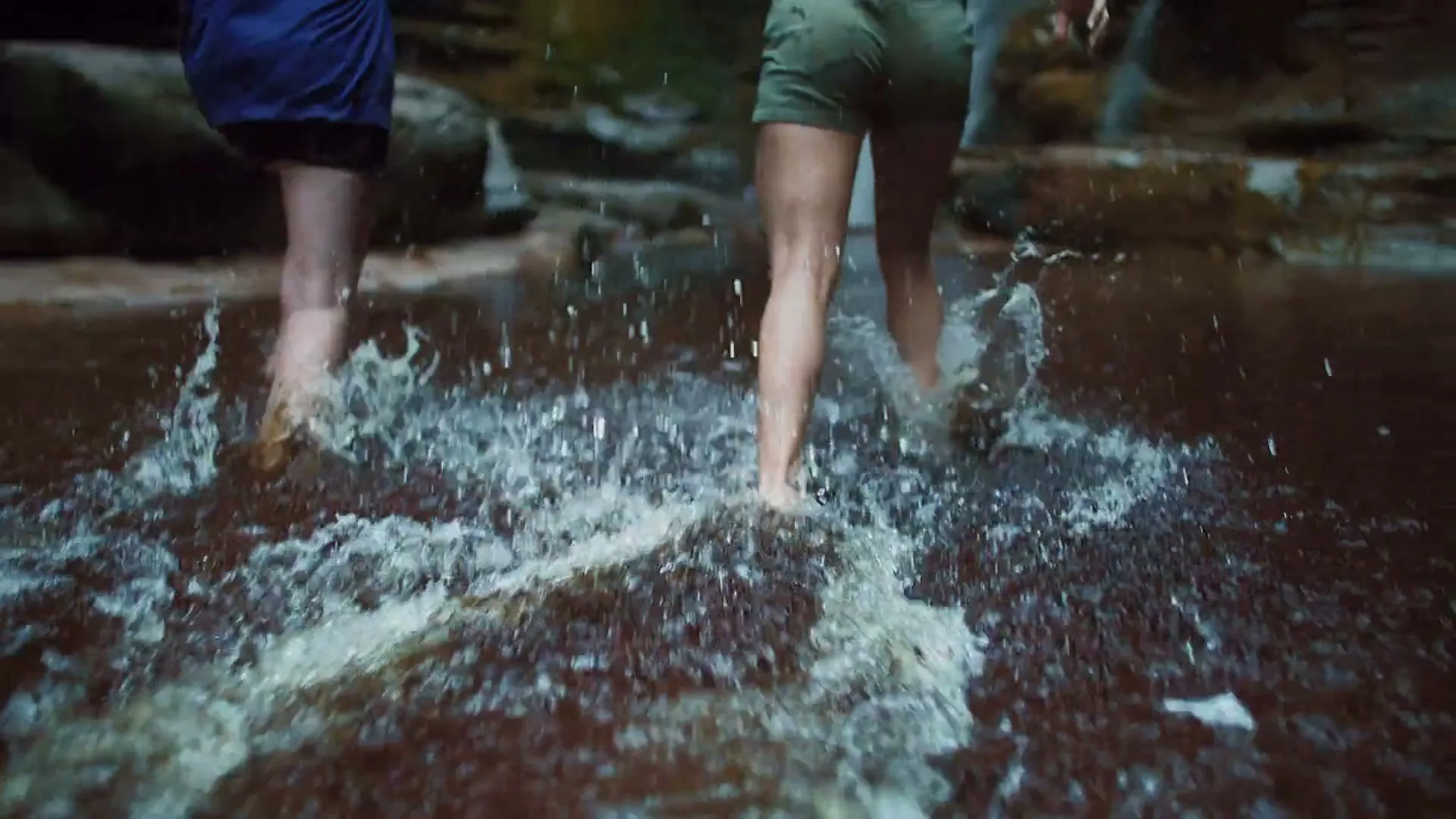 Two young women run barefoot through shallow water, splashing as they approach a waterfall in a rocky canyon.