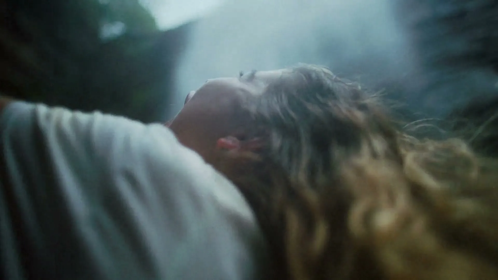 A woman with long, wavy hair and a white shirt looks up with her mouth open, as if screaming or singing, with light shining down on her.