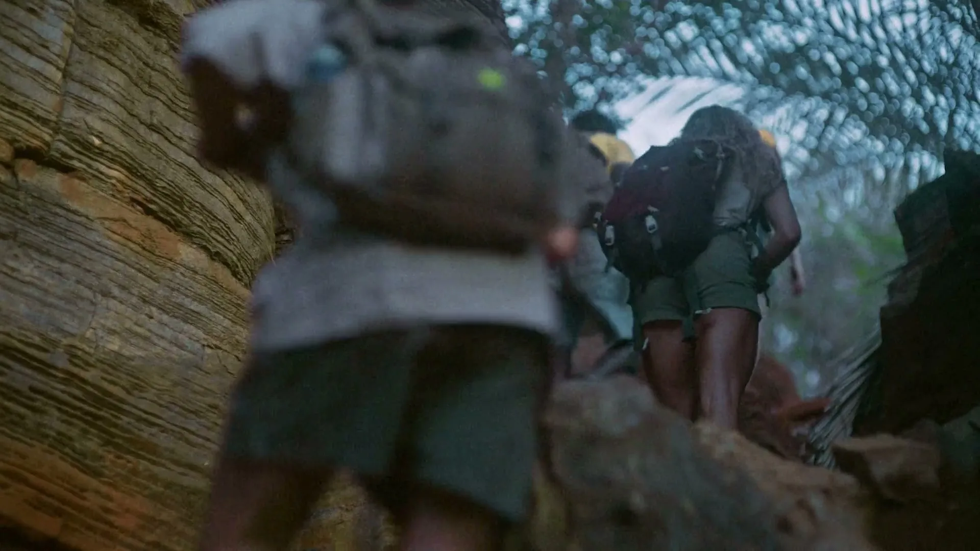 A group of friends, wearing hiking gear, walk through a rocky, jungle-like path, with one person in the foreground blurred.