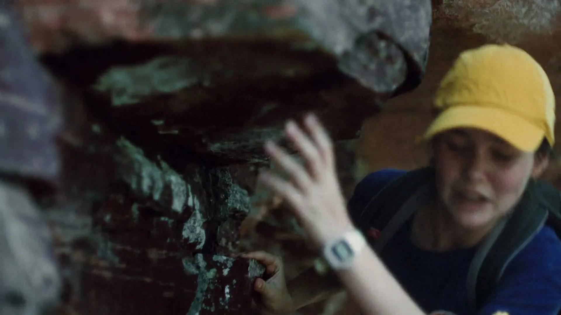 A young woman in a yellow cap and backpack carefully climbs a rocky cave wall, her face showing concentration.