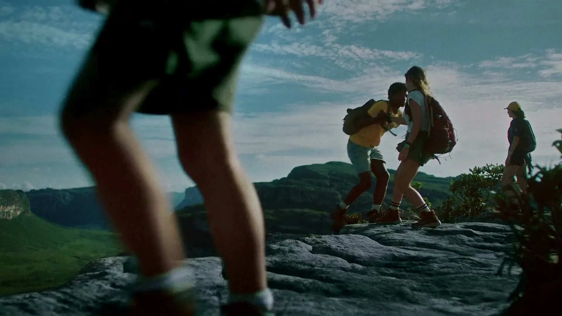 A diverse group of young adults, wearing hiking gear, help each other climb a rocky mountain peak with a vast landscape in the background.