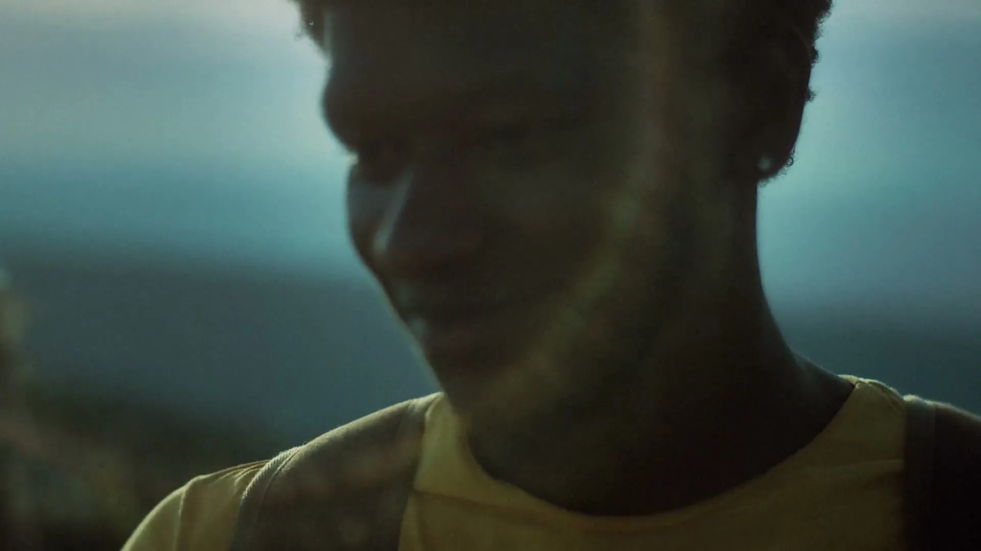 A young Black man in a yellow shirt smiles shyly, looking down and to the side, with a blurred person in the foreground.