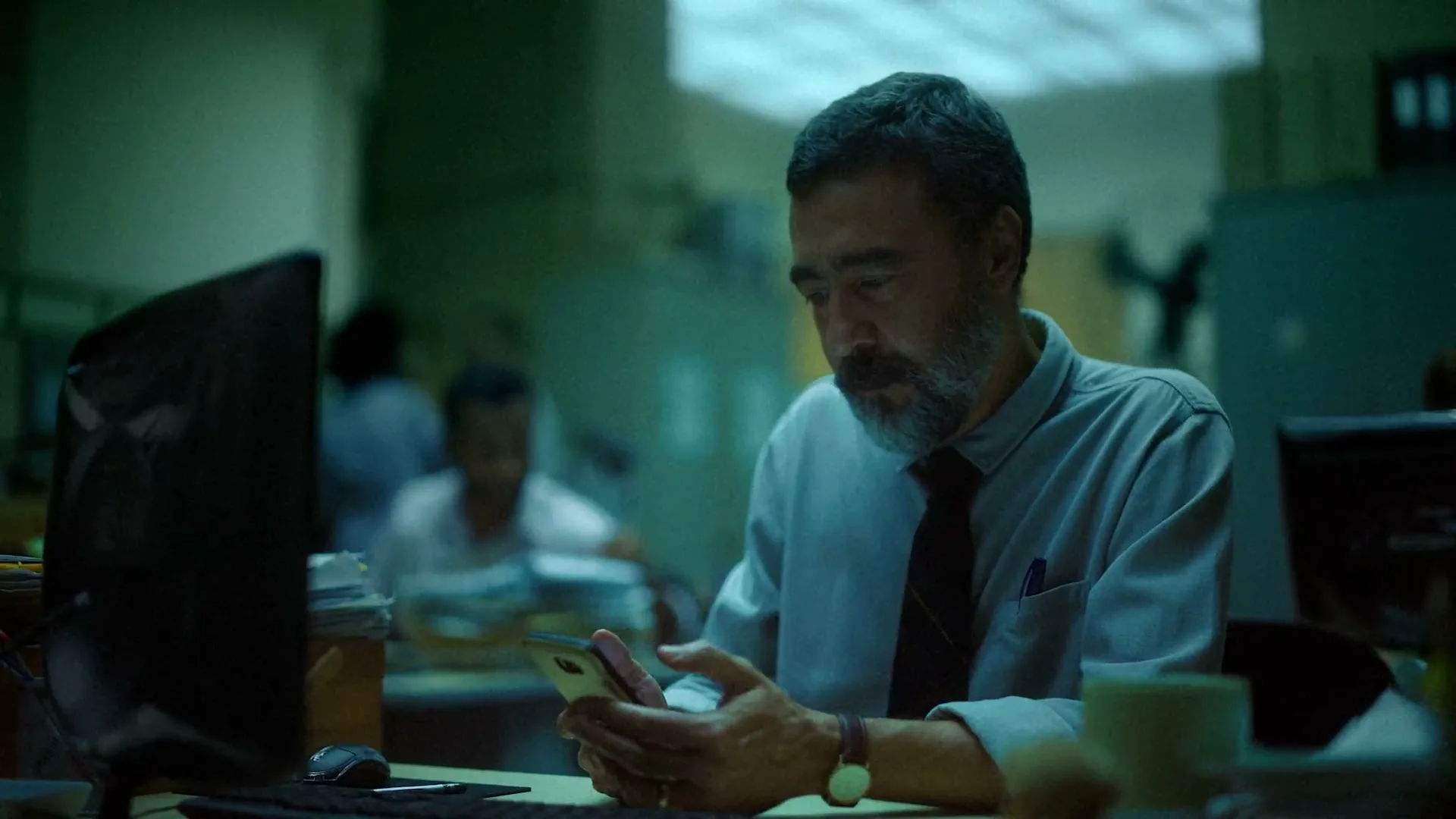 A middle-aged man with a beard, wearing a light blue shirt and dark tie, sits at a desk in a dimly lit office, looking at his phone and smiling.