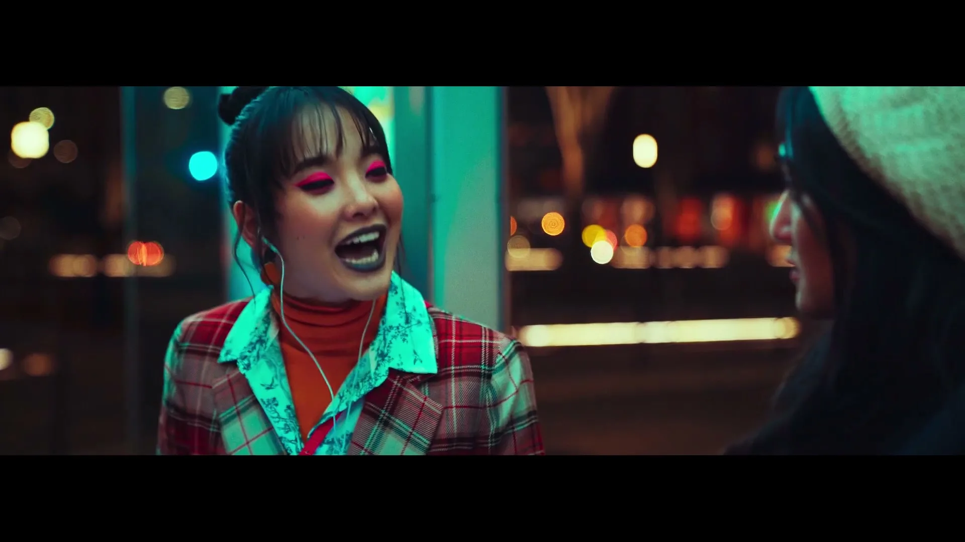 Two young women, one with bright pink eyeshadow and a plaid jacket, talk to each other on a city street at night.