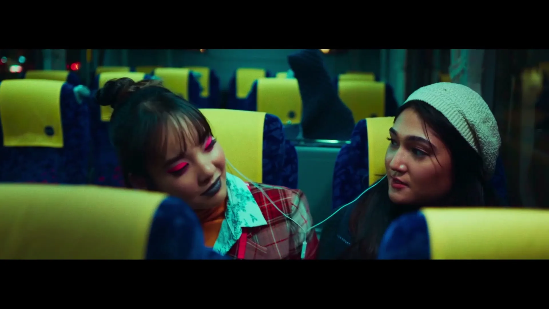 Two young women with earbuds share music on a bus at night, one with bright pink eyeshadow and the other in a beanie.