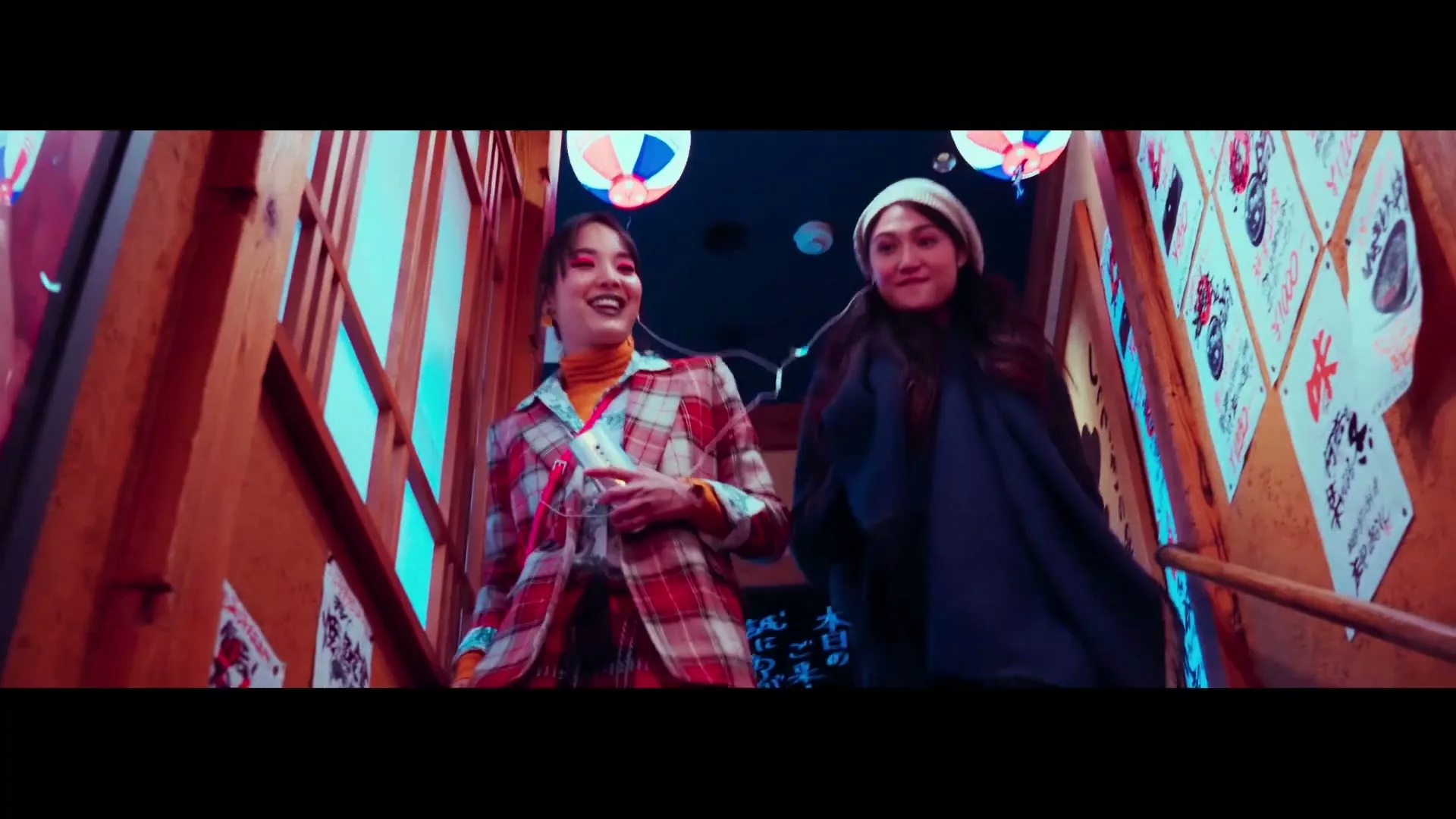 Two young women, one in a plaid jacket and the other in a dark coat, walk down a brightly lit wooden staircase in a Japanese-style establishment.