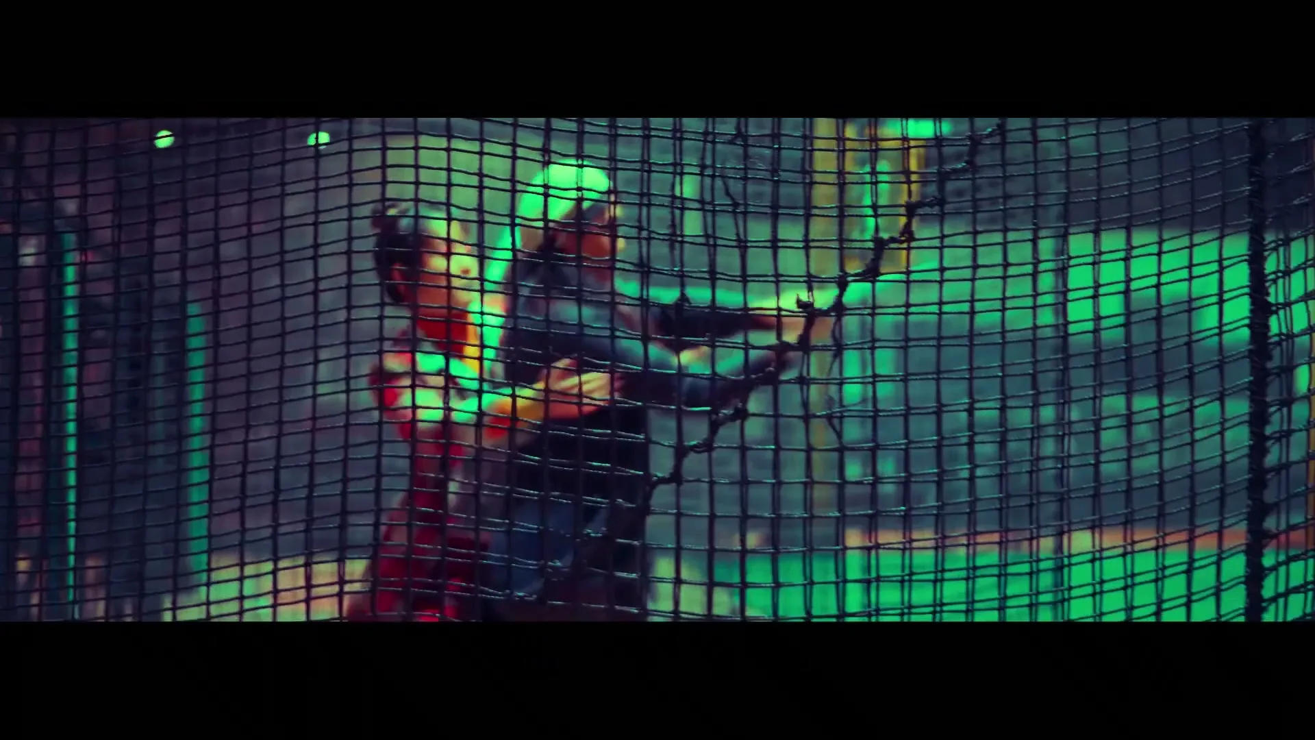 Two young men, one in a red shirt and one in a dark shirt, play paintball behind a net in a dark, neon-lit arena.