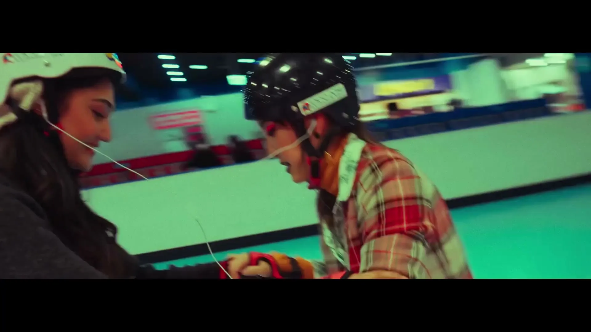 Two young women, one in a plaid shirt and the other in a dark jacket, wear helmets and roller skate together in an indoor rink.