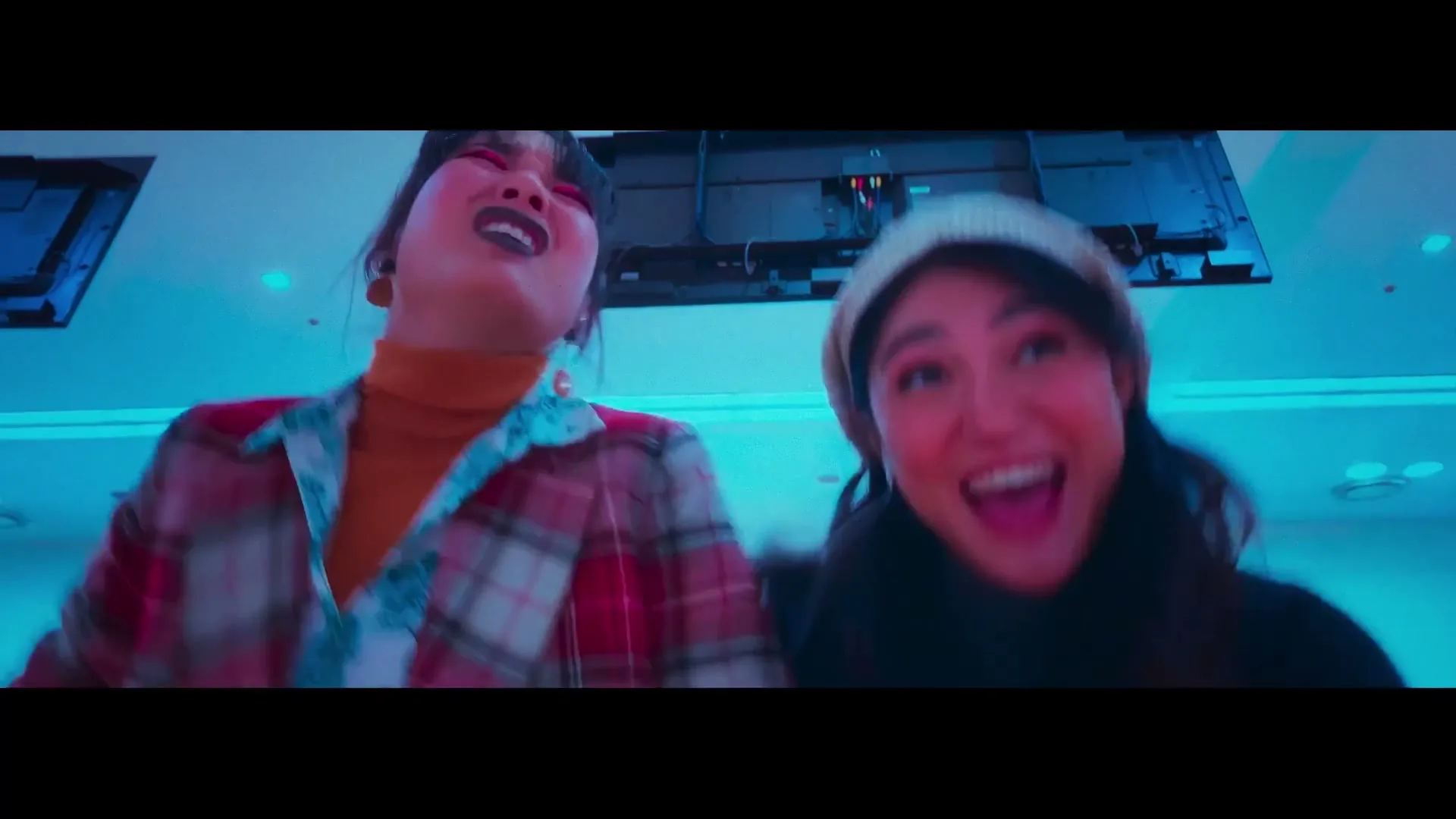 Two young women, one with dark lipstick and a plaid jacket, the other in a beanie, are seen from a low angle, screaming and laughing in a brightly lit, blue-hued room.