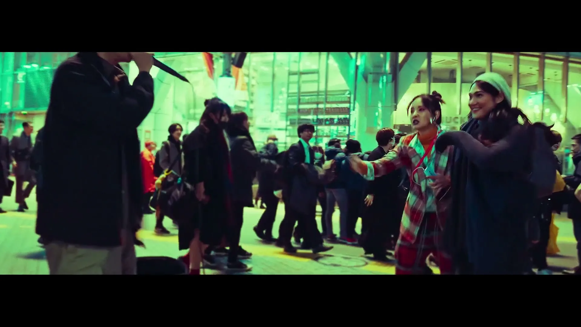 A diverse group of people, including a man singing into a microphone and a woman dancing in a plaid suit, gather on a busy city street at night.