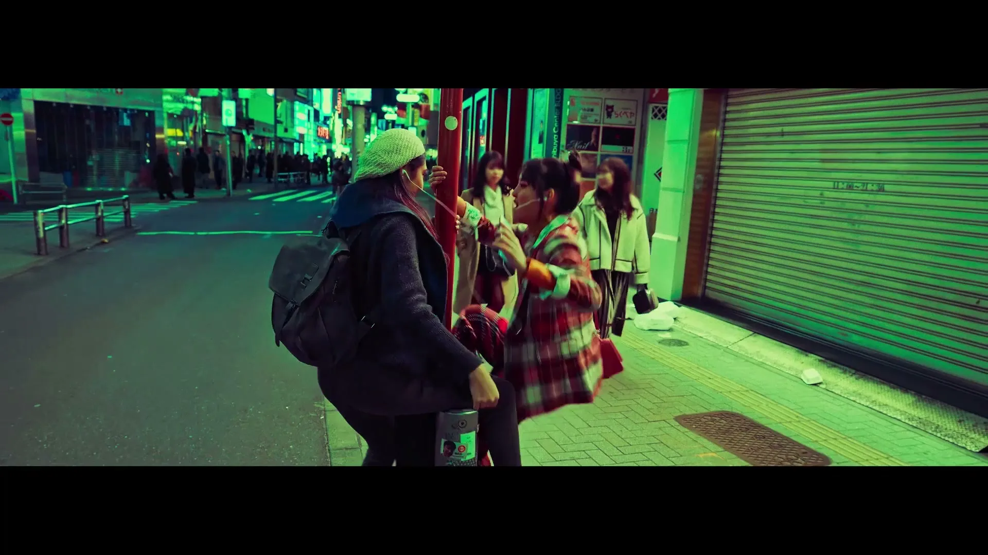 Two young women share earbuds and dance playfully on a city street at night, surrounded by neon lights and closed shops.