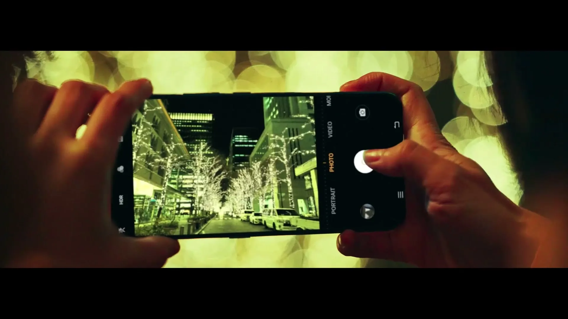 A person holds a smartphone horizontally, taking a photo of a city street at night, with bokeh lights in the background.