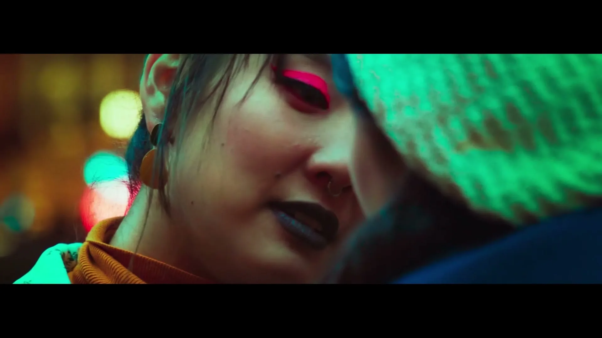 A young woman with bright pink eyeshadow and a nose ring looks at another person, their face partially obscured, in a vibrant, blurry urban setting at night.