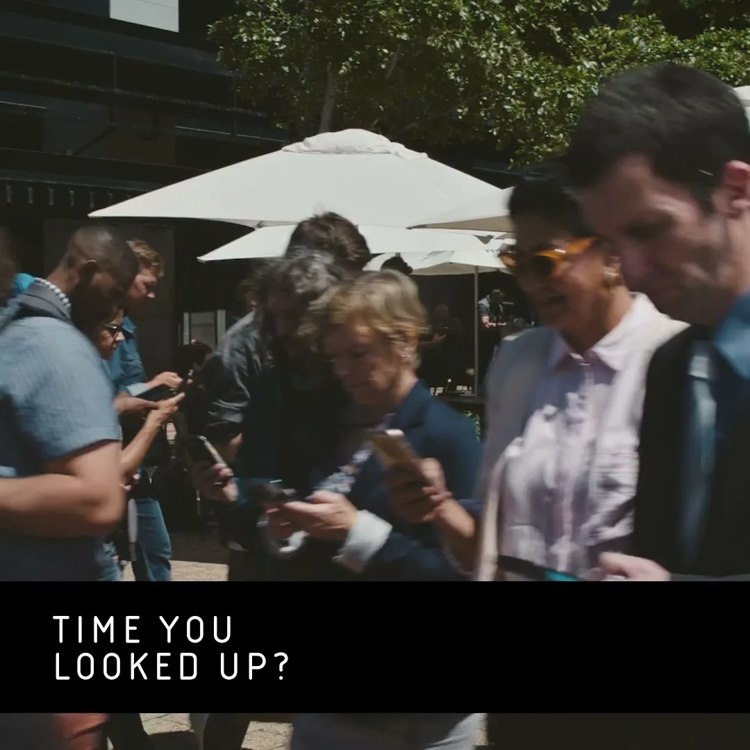 A diverse crowd of people walk past the camera, all looking down at their phones, in an outdoor urban setting.