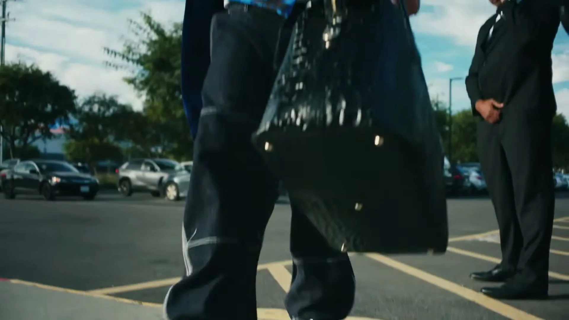 A man in a suit and blue vest, holding sunglasses, stands in a parking lot and points, with a blue sky and clouds in the background.