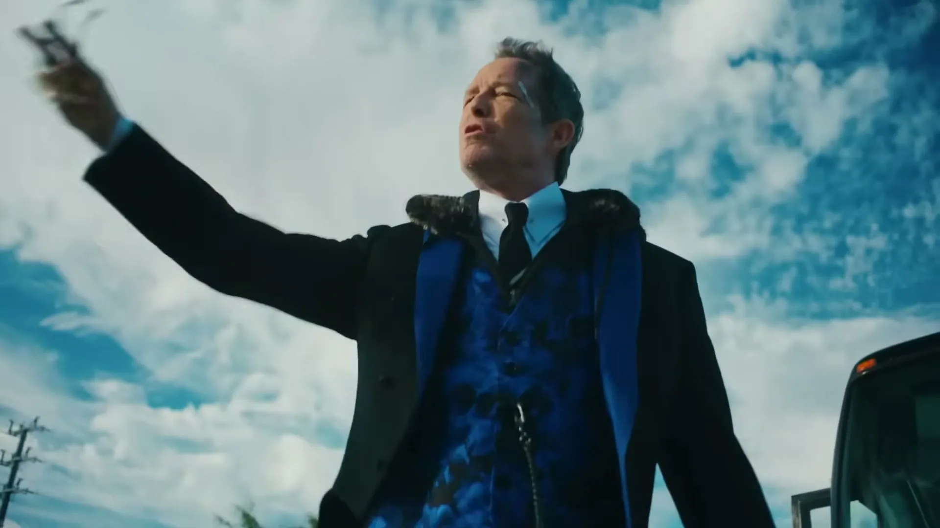 A man in a suit and blue vest stands outside, looking up and throwing something into the air against a cloudy sky.