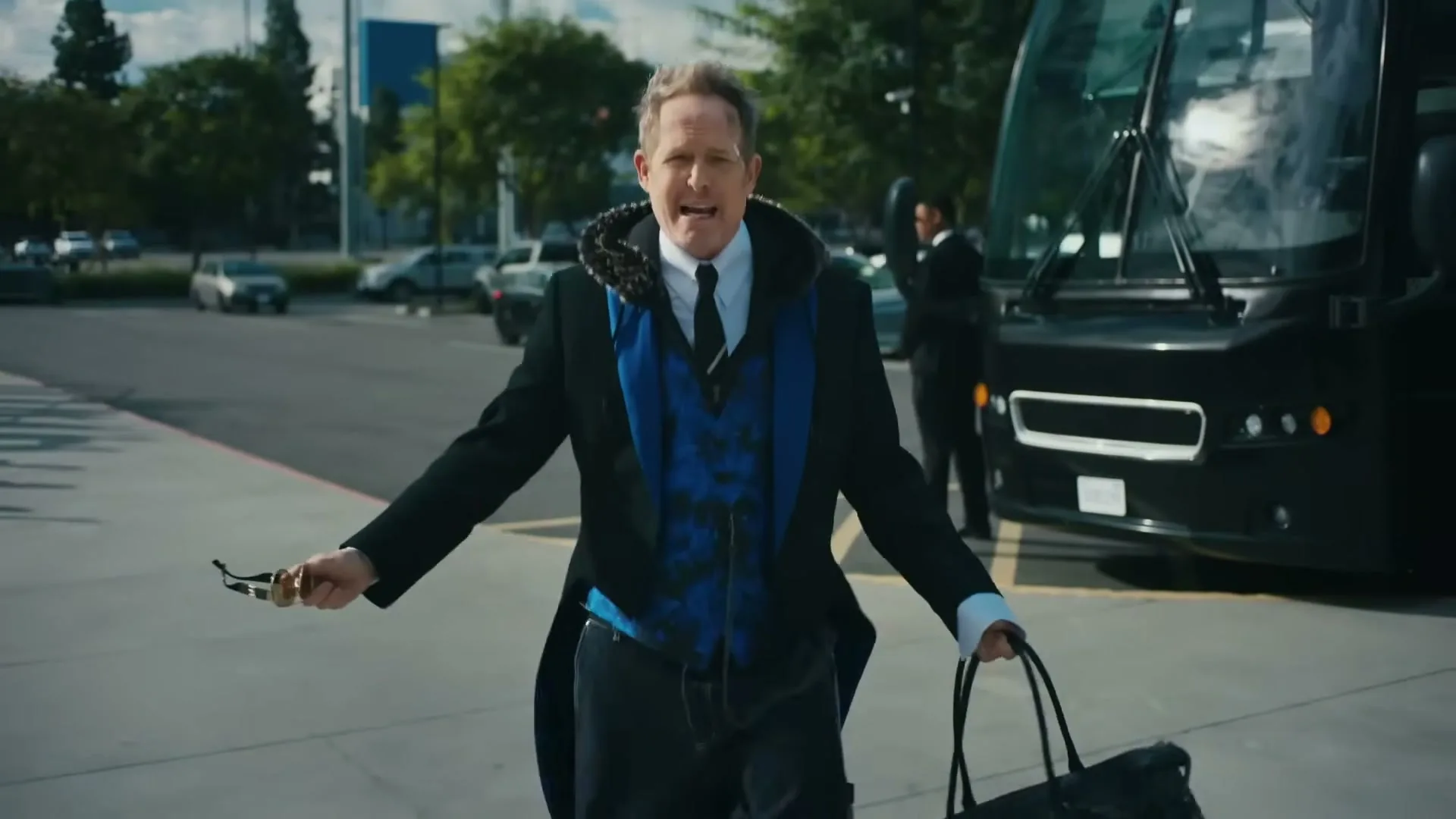 A man in a black coat and blue vest walks towards the camera, holding a bag and glasses, with a bus and other cars in the background.