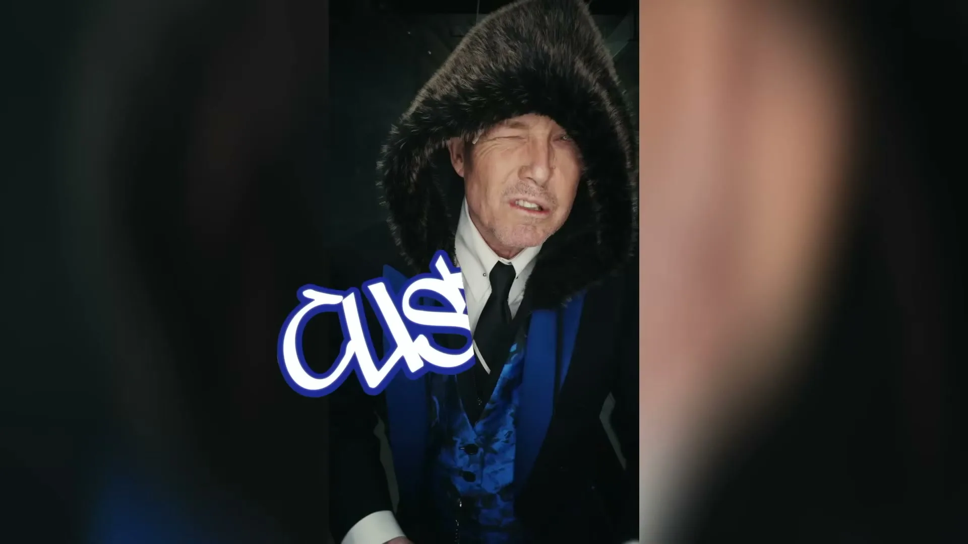 A man in a fur-lined hood and suit winks and gestures with his fingers, as the word "CUSTOM" appears on screen in a dark setting.
