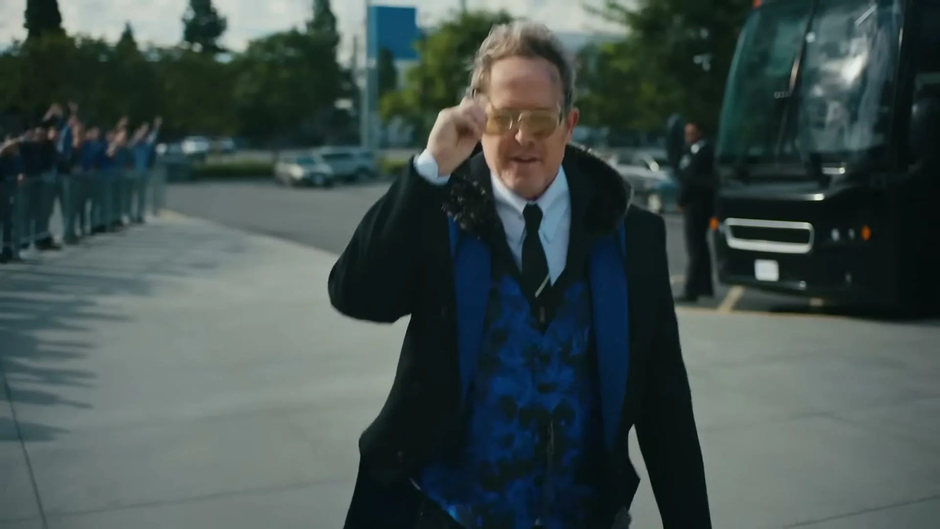 A man in a black coat and blue vest walks towards the camera, putting on sunglasses, with a bus and people in the background.