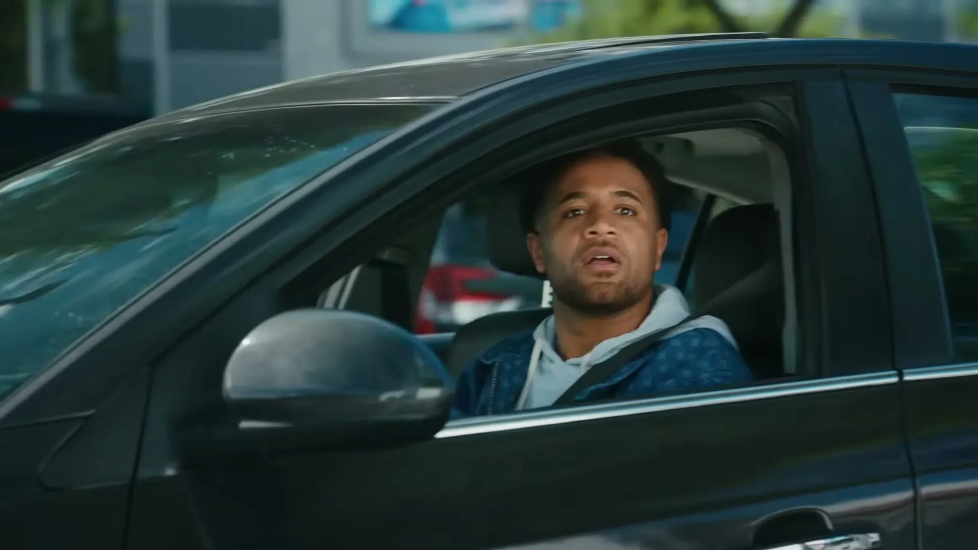 A man with a beard and a blue jacket is driving a black car, looking surprised and slightly scared.