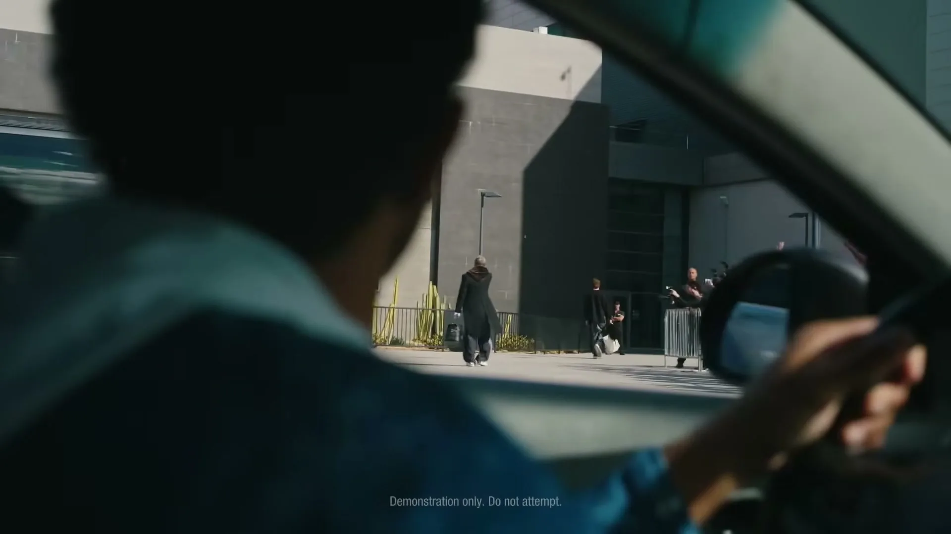 A driver in a blue hoodie looks over their shoulder at a man in a black coat walking away from a building, as a bus passes by.
