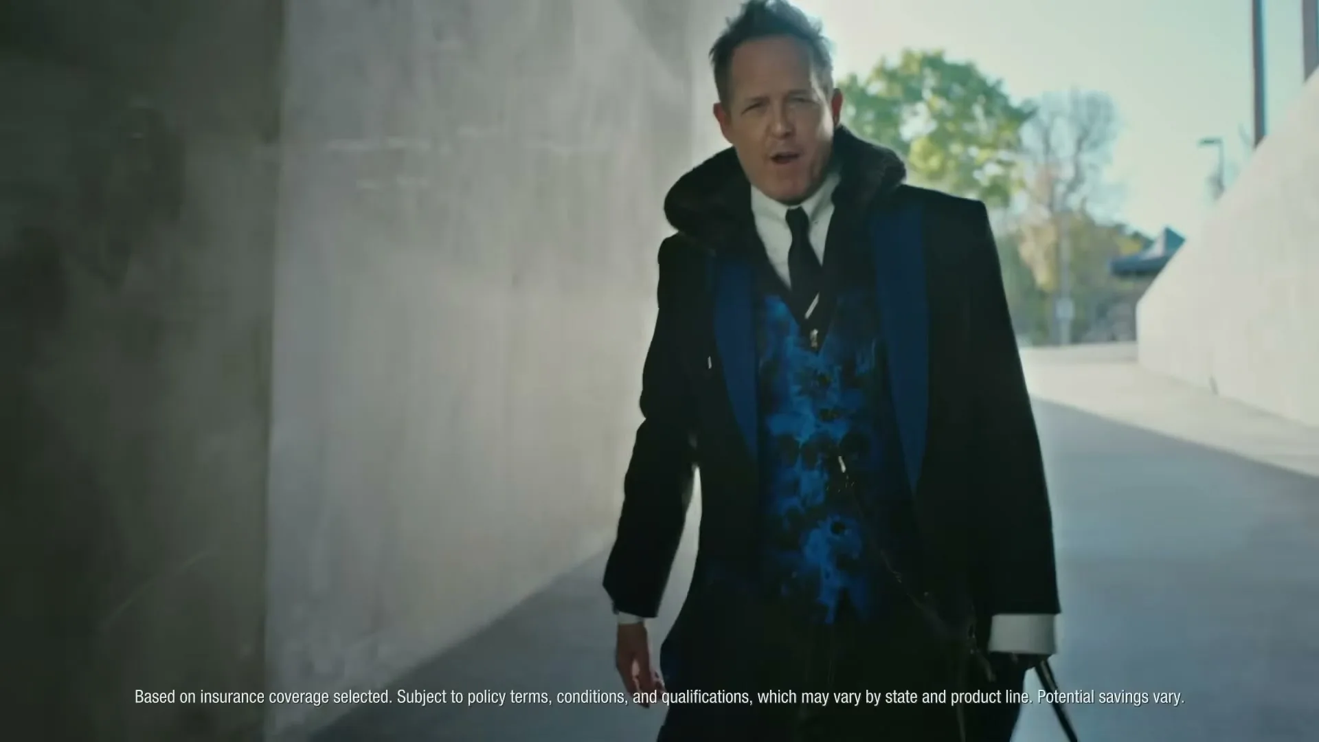 A man in a black suit with a blue vest walks towards the camera in a concrete underpass, speaking directly to the viewer.