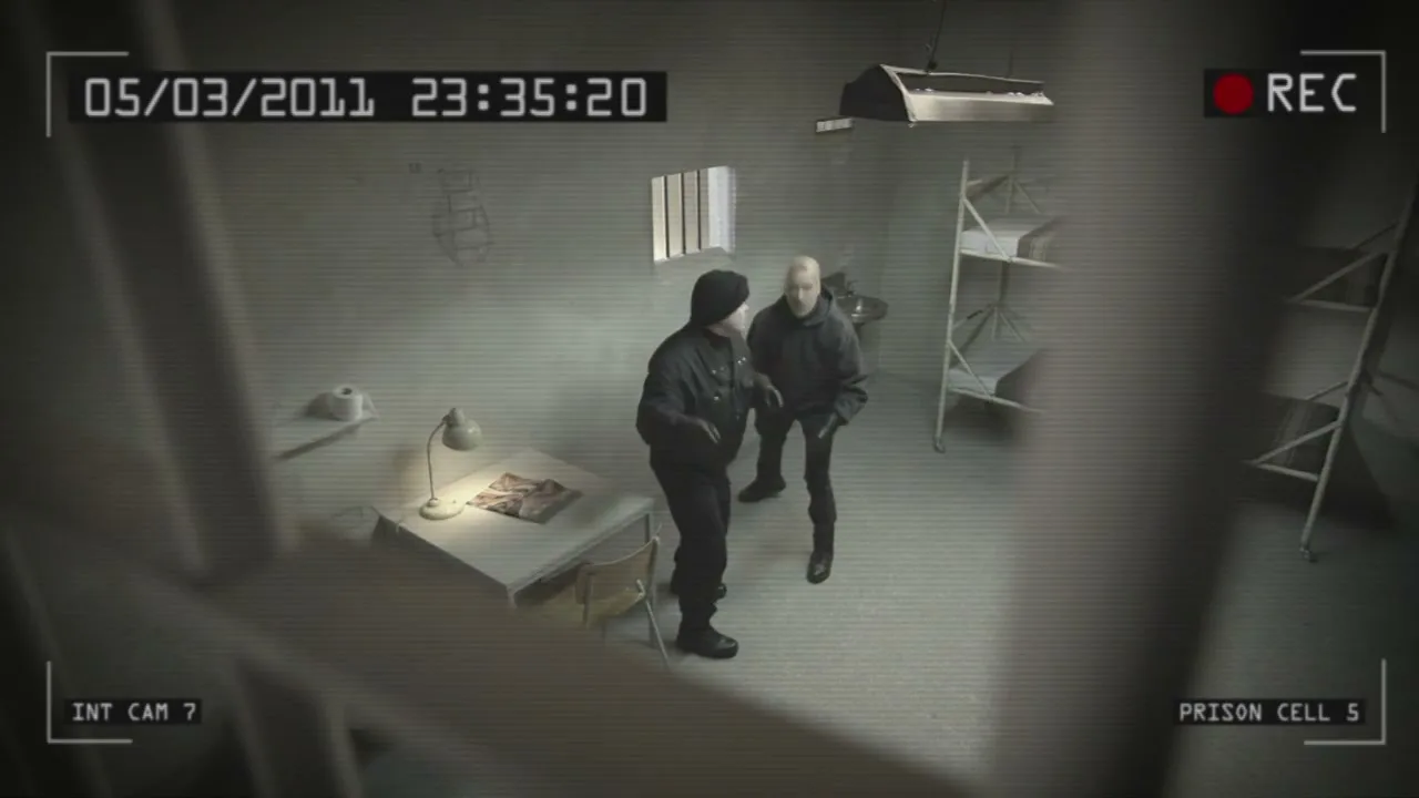 Two men in dark clothing stand in a prison cell, while other prisoners move around them, all seen through a security camera lens.