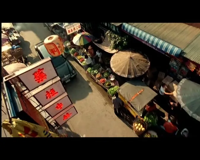 An overhead shot of a bustling street market with vendors, umbrellas, and vehicles, as the camera quickly pans down the street.