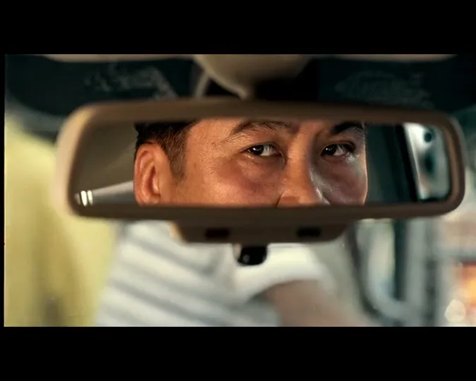 A man's eyes are reflected in a car's rearview mirror, looking intently forward while driving during the day.