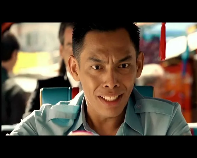 A male tuk-tuk driver in a light blue shirt looks back at the camera with a wide, excited grin, while a man in a suit smiles behind him in a bustling street.